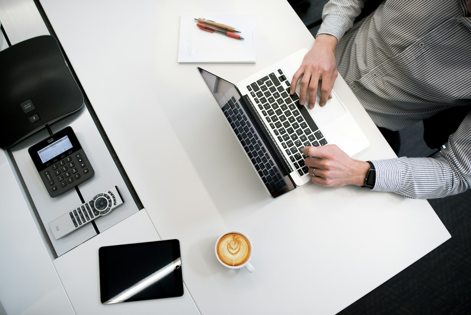 Persoon aan een wit bureau met laptop, telefoon en notitieblok